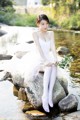 A woman in a white dress sitting on a rock by the water.