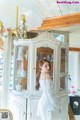 A woman in a white dress standing in front of a china cabinet.